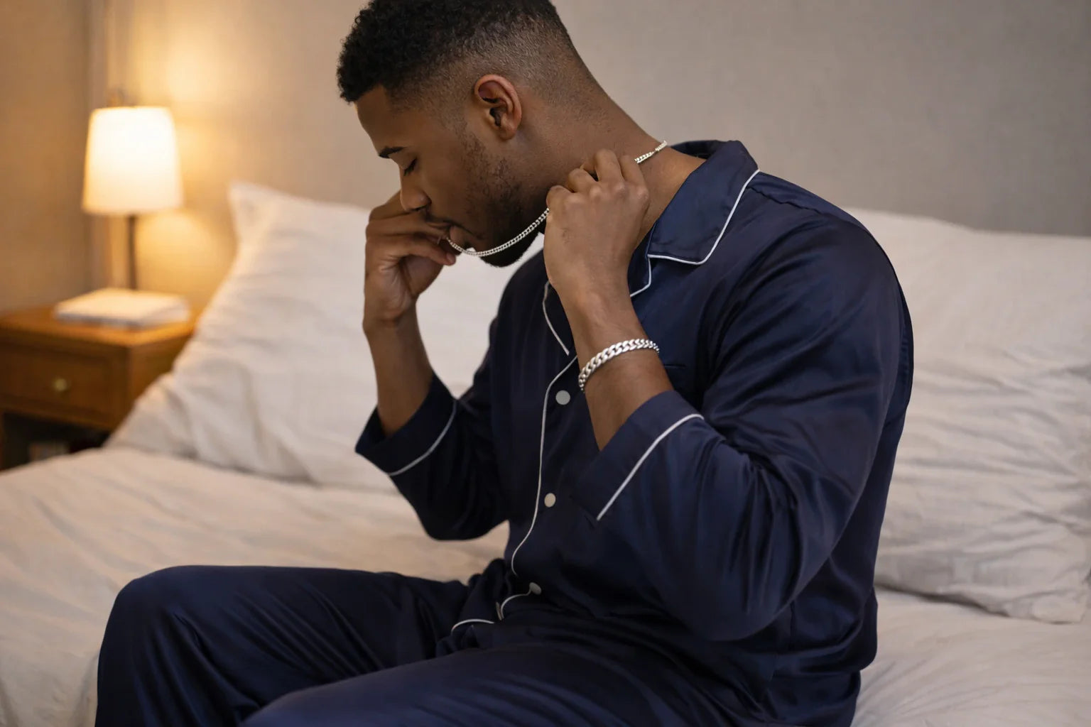 Photo – a man sitting on the edge of a bed, gently taking off a sterling silver chain while wearing a single silver bracelet, showing a natural nighttime routine and reflecting can you sleep with
